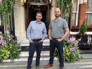Two men stand in front of a building entrance adorned with lush greenery and flowers. Both are casually dressed, one in a light blue striped shirt and the other in a gray polo.