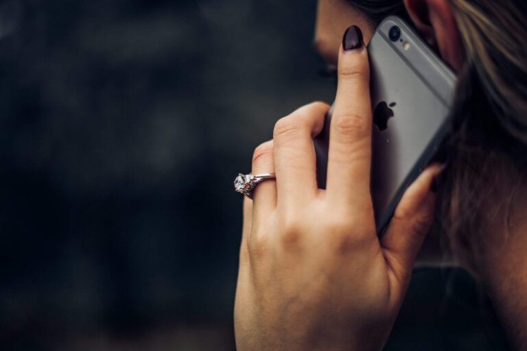 A close-up shot of a person holding an iPhone to their ear, indicating a phone conversation. The person has dark, polished nails and wears a large, sparkling diamond ring on their ring finger.