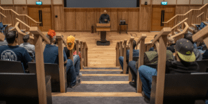 A CovertSwarm Academy intake session in a modern lecture hall with wooden paneling, featuring a speaker at a podium addressing academy candidates.