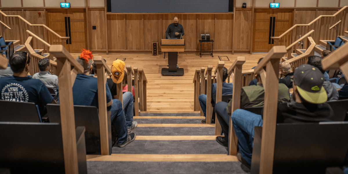 A CovertSwarm Academy intake session in a modern lecture hall with wooden paneling, featuring a speaker at a podium addressing academy candidates.