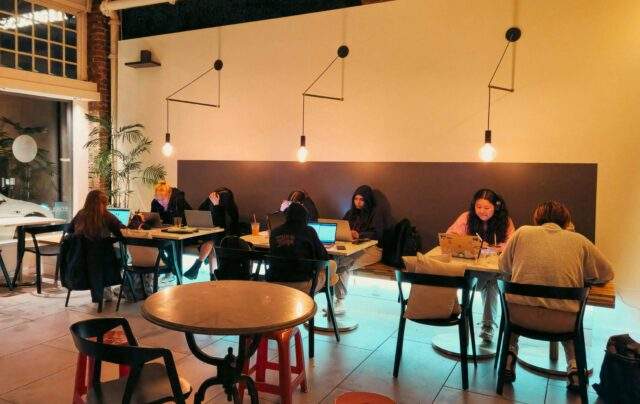 People working remotely on laptops in a café environment, seated at separate tables.