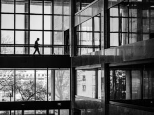 Black-and-white photo of a silhouetted person walking through a glass-and-marble office atrium, unaware of what's exposed just out of sight.