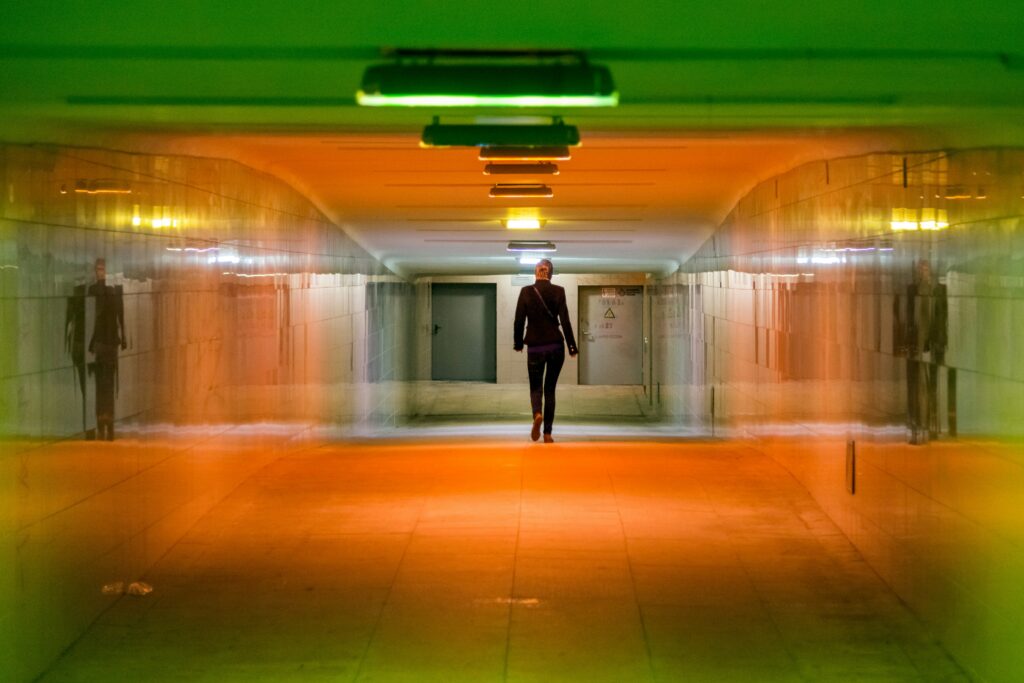 A lone figure walking through a long illuminated corridor, a motion sensor above them glowing green, the alert never coming.