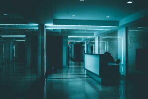 A dim, teal-lit office corridor with a lone figure at a reception desk and a distant exit sign, evoking the physical intrusion staff walked past without noticing.