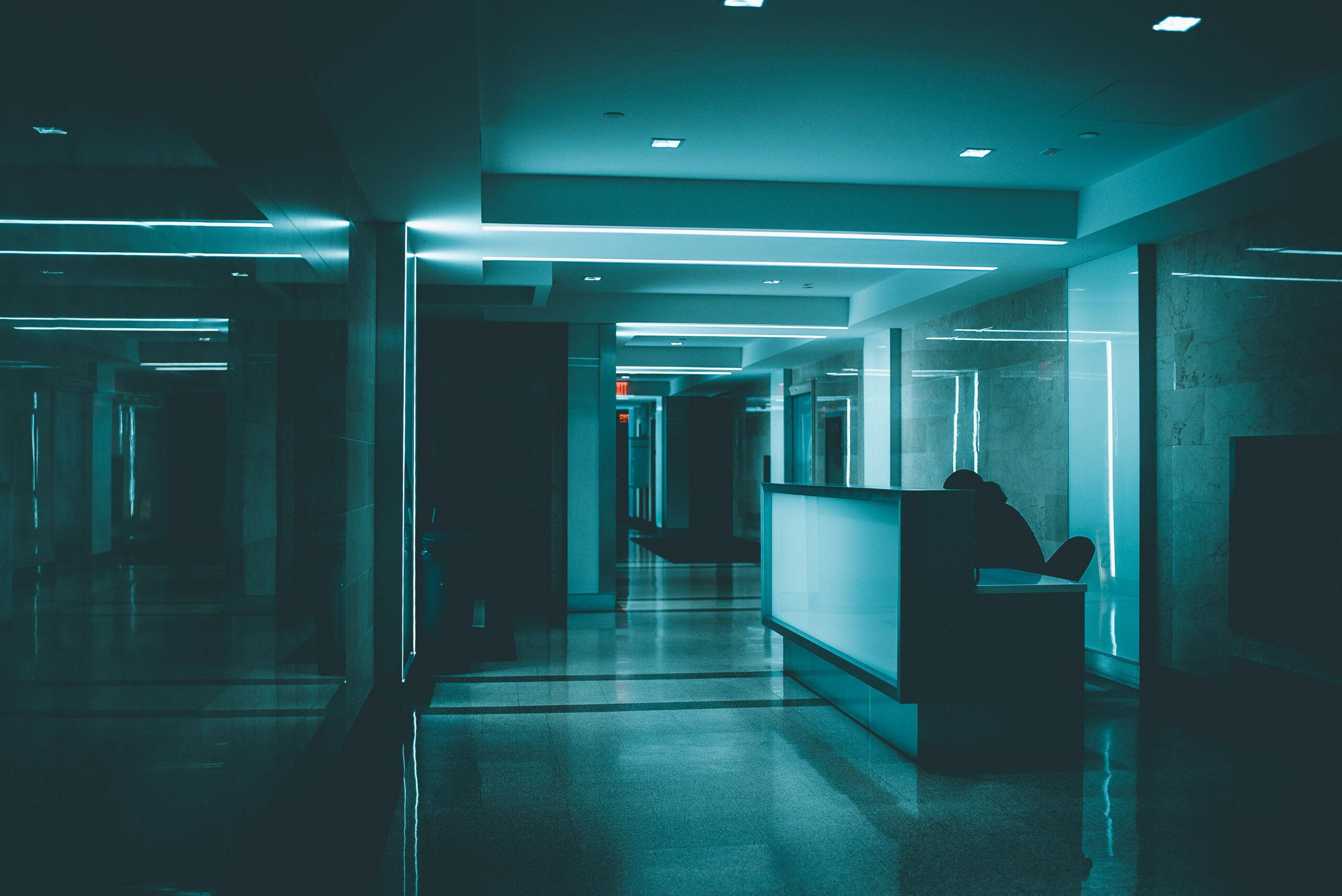 A dim, teal-lit office corridor with a lone figure at a reception desk and a distant exit sign, evoking the physical intrusion staff walked past without noticing.