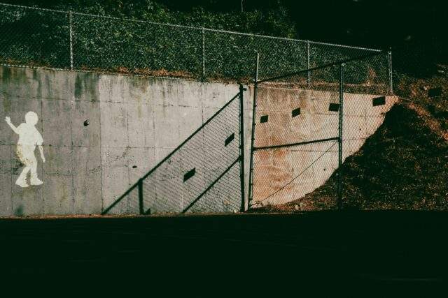 Stencil silhouette of a child walking along a concrete wall beside a gap in a chain-link fence, symbolizing a gap in AI agent governance and containment boundaries