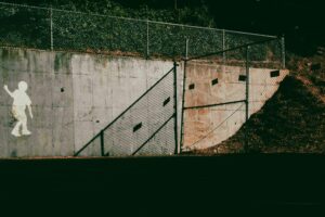 Stencil silhouette of a child walking along a concrete wall beside a gap in a chain-link fence, symbolizing a gap in AI agent governance and containment boundaries