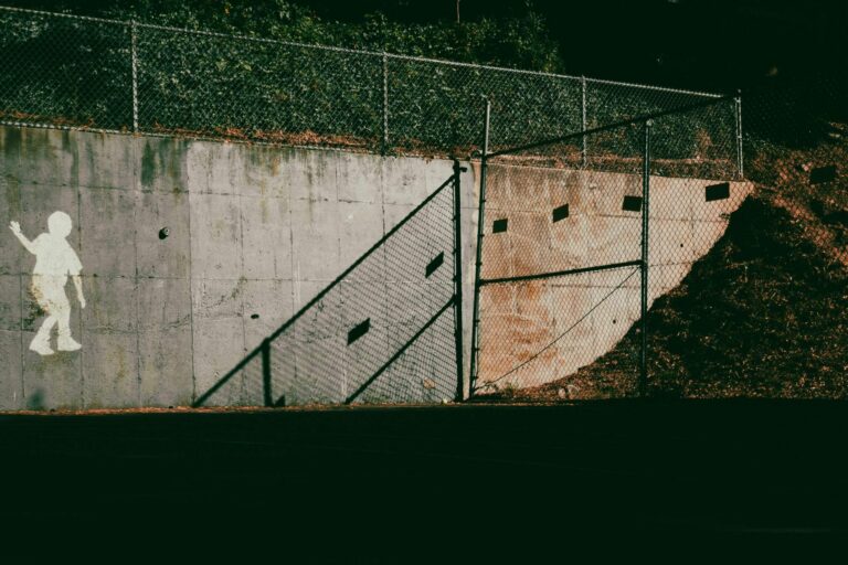 Stencil silhouette of a child walking along a concrete wall beside a gap in a chain-link fence, symbolizing a gap in AI agent governance and containment boundaries