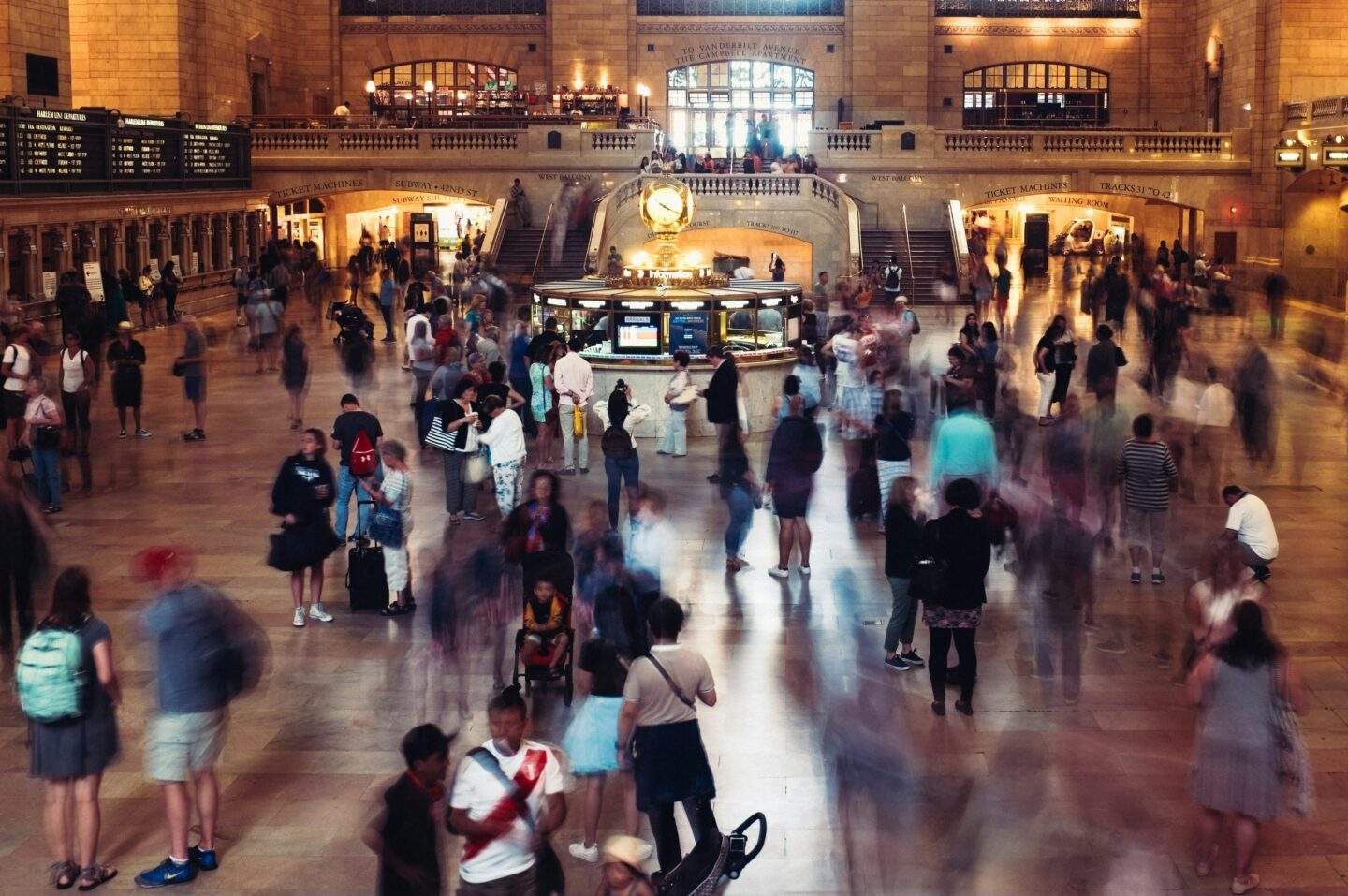 Long-exposure shot of a crowded station concourse, with a static, illuminated information booth at the center while blurred figures move past in every direction.