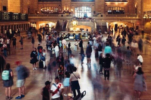 Long-exposure shot of a crowded station concourse, with a static, illuminated information booth at the center while blurred figures move past in every direction.