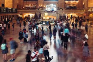 Long-exposure shot of a crowded station concourse, with a static, illuminated information booth at the center while blurred figures move past in every direction.