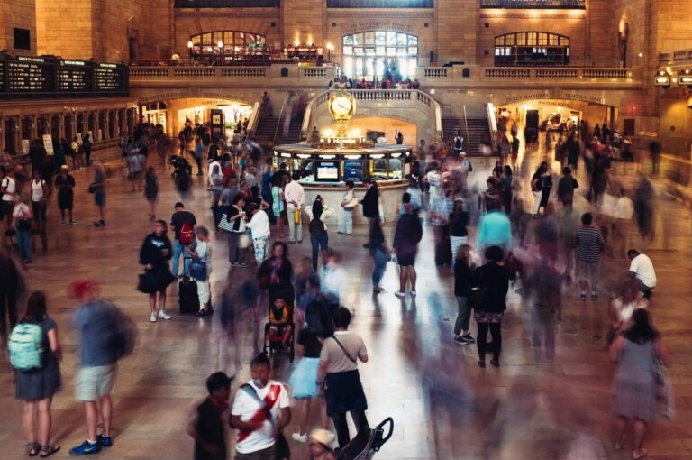Long-exposure shot of a crowded station concourse, with a static, illuminated information booth at the center while blurred figures move past in every direction.