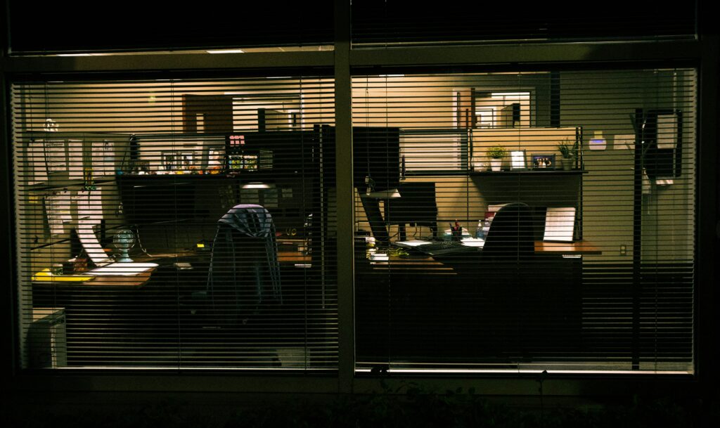 An office seen through closed blinds at night, desks lit but empty, evoking an unauthorized presence with a view into systems nobody's watching.