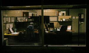 An office seen through closed blinds at night, desks lit but empty, evoking an unauthorized presence with a view into systems nobody's watching.