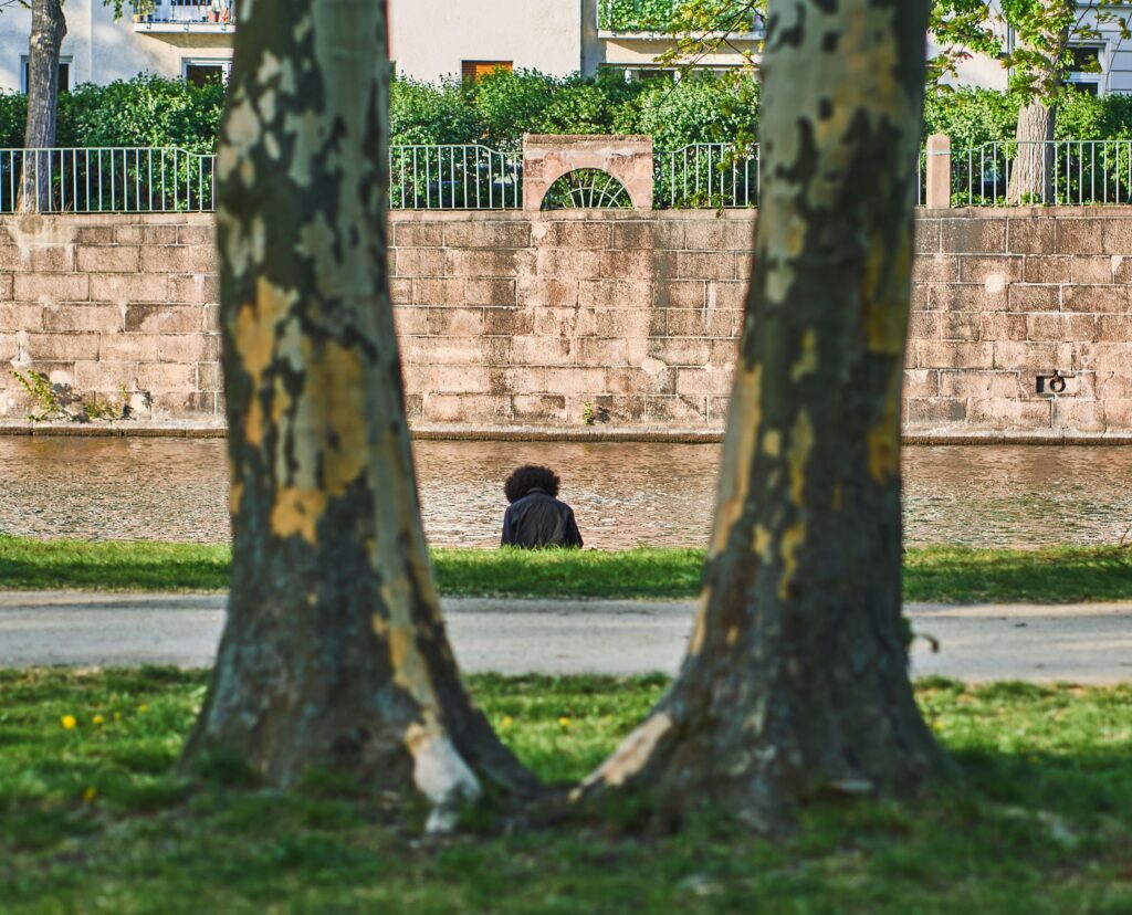 A solitary figure sits by a canal wall, viewed unnoticed from a hidden vantage point between two tree trunks, evoking an assumed breach attacker moving undetected.