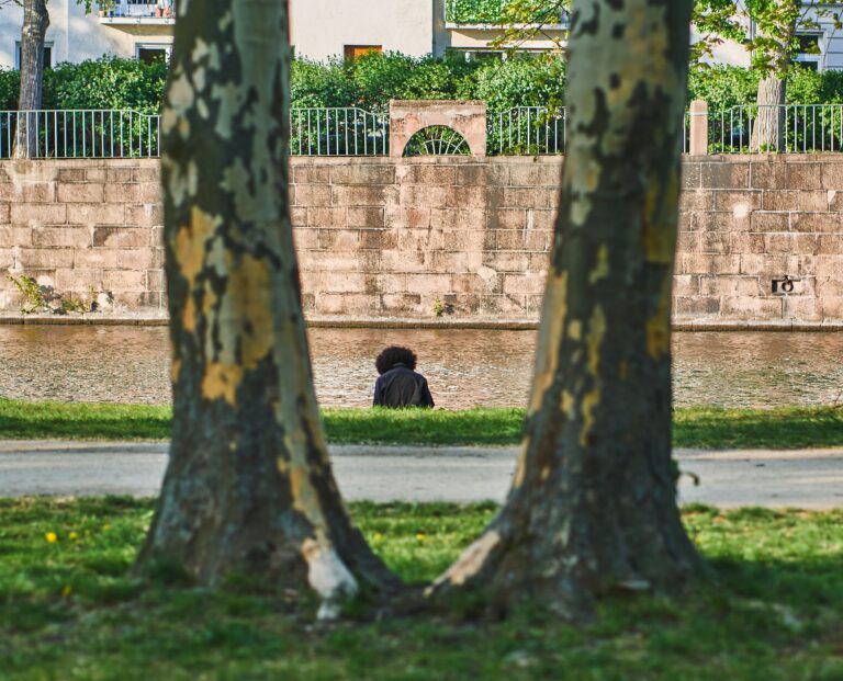 A solitary figure sits by a canal wall, viewed unnoticed from a hidden vantage point between two tree trunks, evoking an assumed breach attacker moving undetected.