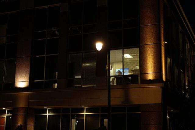 A single lit office window glows in an otherwise dark building at night, a monitor visible and unattended inside.