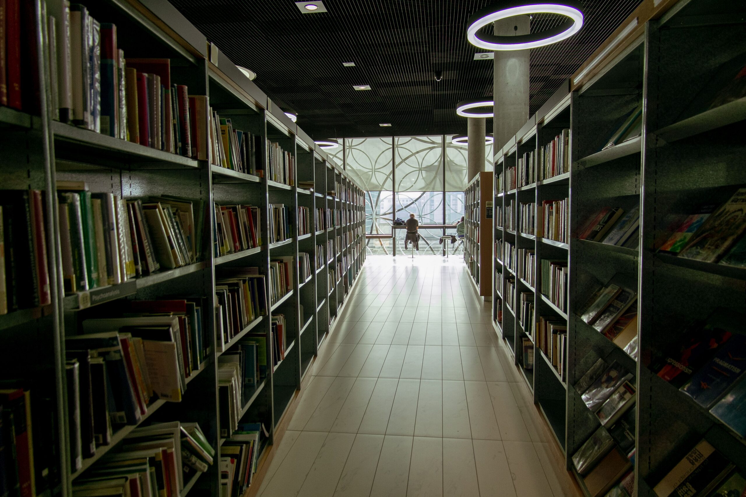 A narrow library aisle between towering bookshelves leading toward a distant lit window, evoking the public data an attack surface mapping exercise can turn into a path to private systems.