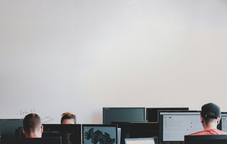 Employees working at their monitors, heads down, unaware their help desk is fielding a vishing attack disguised as a routine password reset.