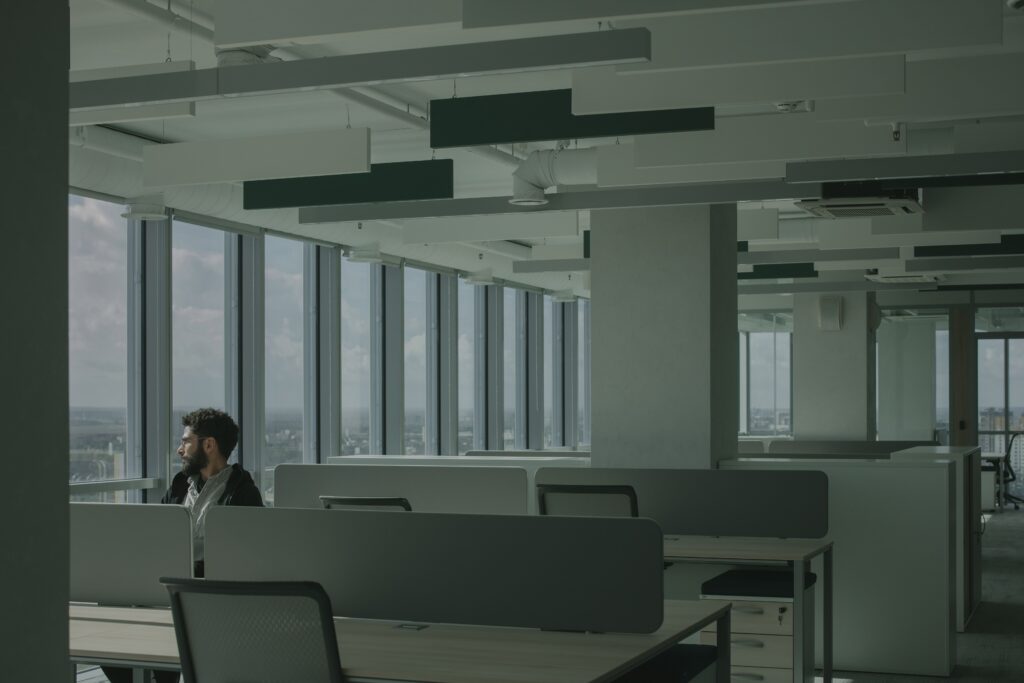 A figure pauses at the edge of a row of empty office desks, looking out over a hazy city skyline through floor-to-ceiling windows.