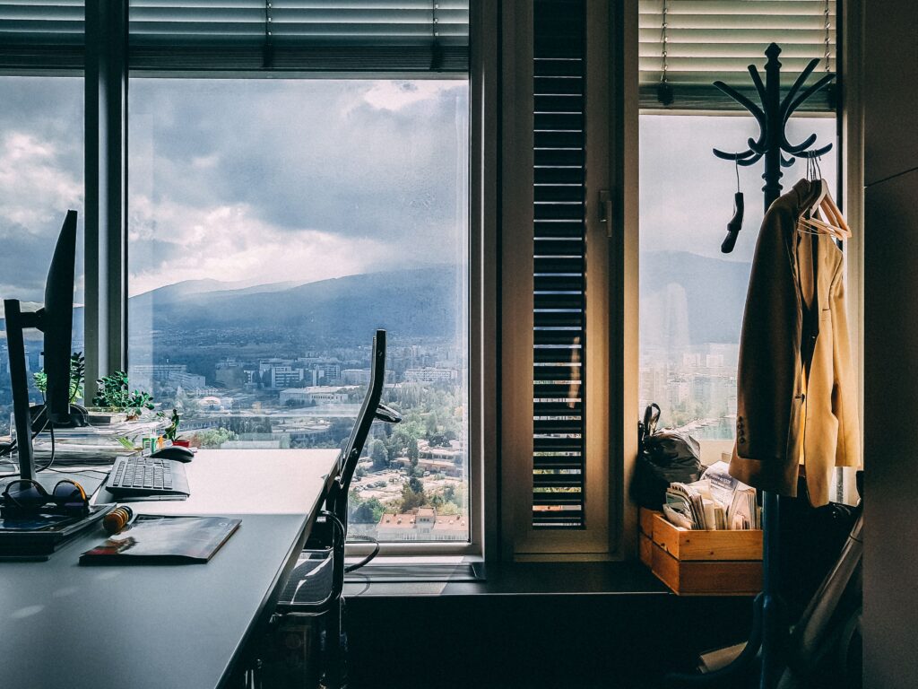 An unattended desk beside a rain-streaked office window, with a coat left hanging and a storm rolling over distant hills.
