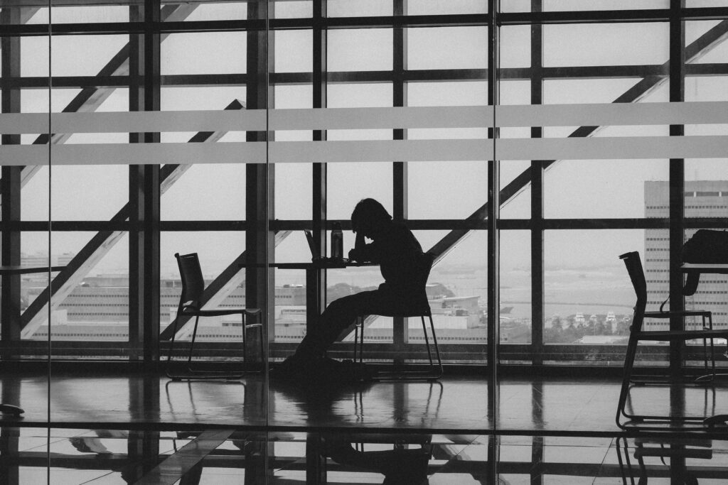 A silhouetted figure works alone at a high-rise desk, dwarfed by a grid of windows overlooking a distant city skyline.